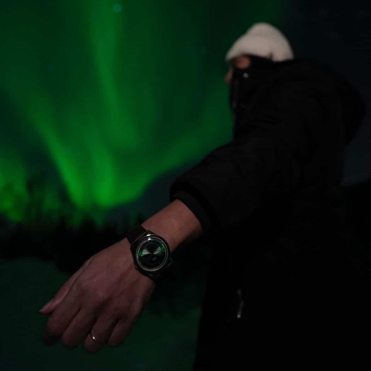 Bilde av arm med en ÁIGI nordlys herreklokke på forran en himmel med nordlys